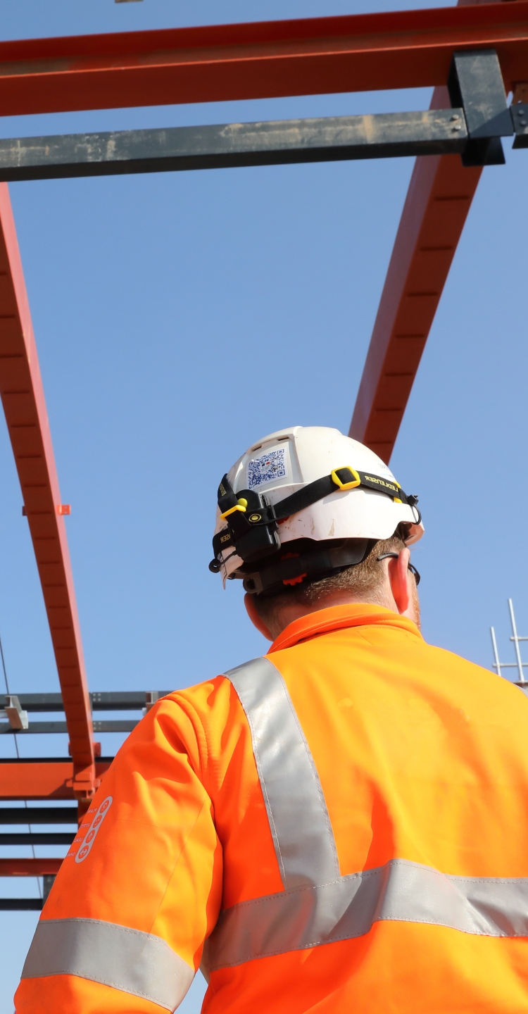 A man stands with his back to the camera. He is on site in high-vis orange clothes looking up at a red steel structure in a clear blue sky.