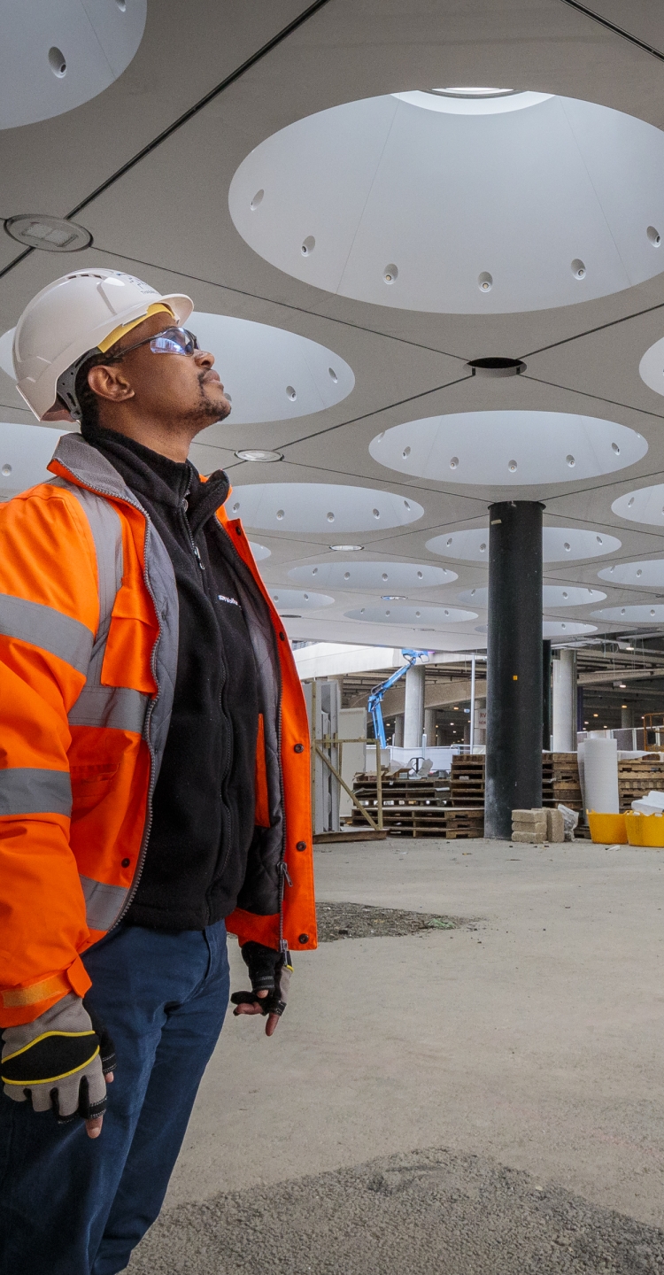 A black man stands on site looking up at a white, modern and sculptural ceiling that is under construction. He wears an orange high-vis jacket and helmet. 