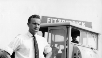 The original founder of the business is pictured in black and white, standing proudly with his hands on his hips, wearing a shirt and tie. In the background is a retro sign with the company name, Fitzpatrick, and a vintage digger. 