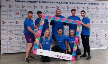 A mixed group of people in charity t-shirts pose next to signage for the Gender Diversity challenge with Women in Rail organisation