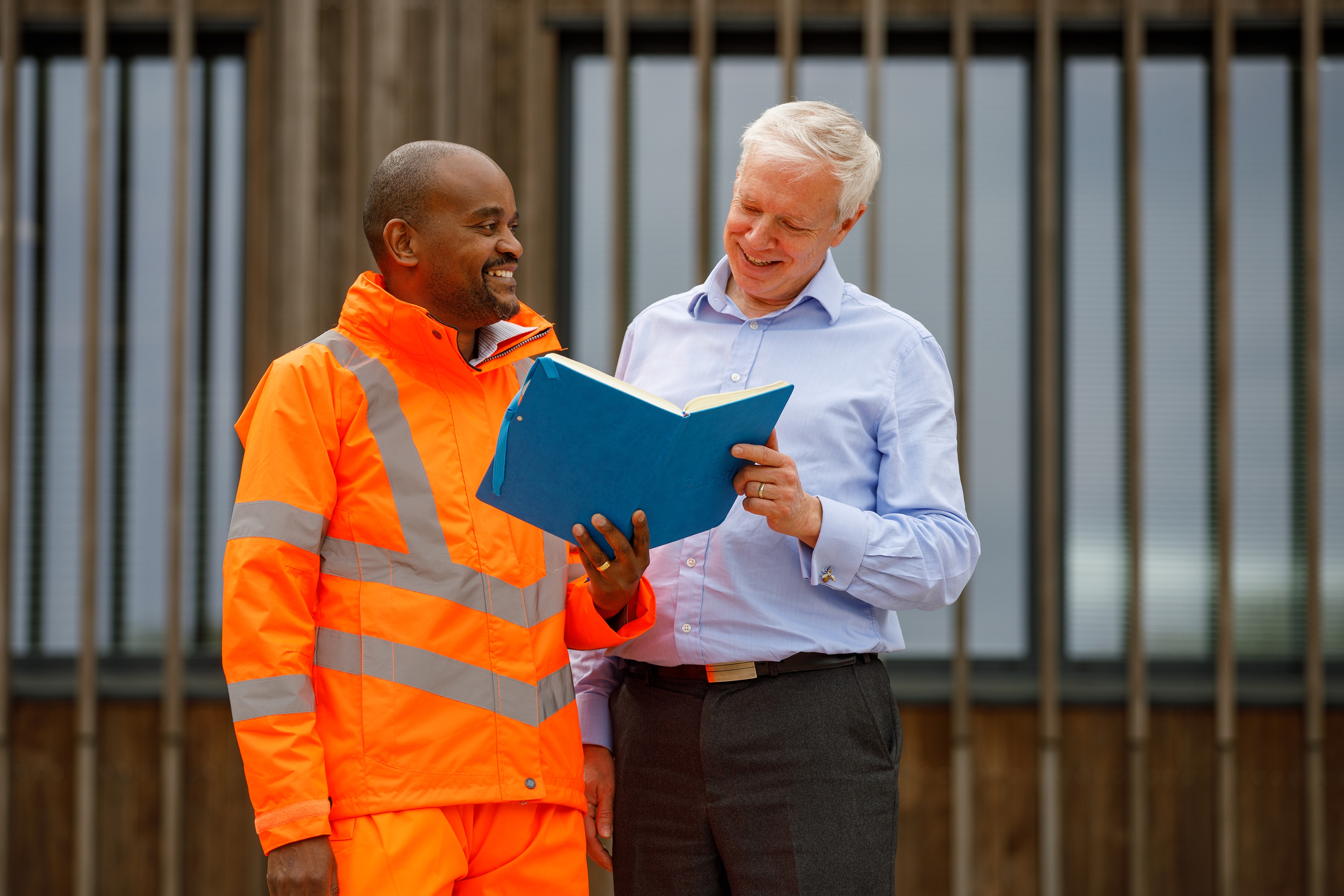This corporate shot show two men stand outside an office. One is in office clothes the other is in high-vis clothing. One holds a book open for the other man to view. The image is diverse.