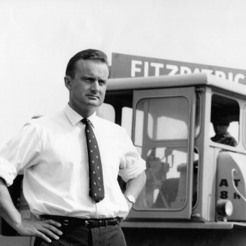The original founder of the business is pictured in black and white, standing proudly with his hands on his hips, wearing a shirt and tie. In the background is a retro sign with the company name, Fitzpatrick, and a vintage digger.